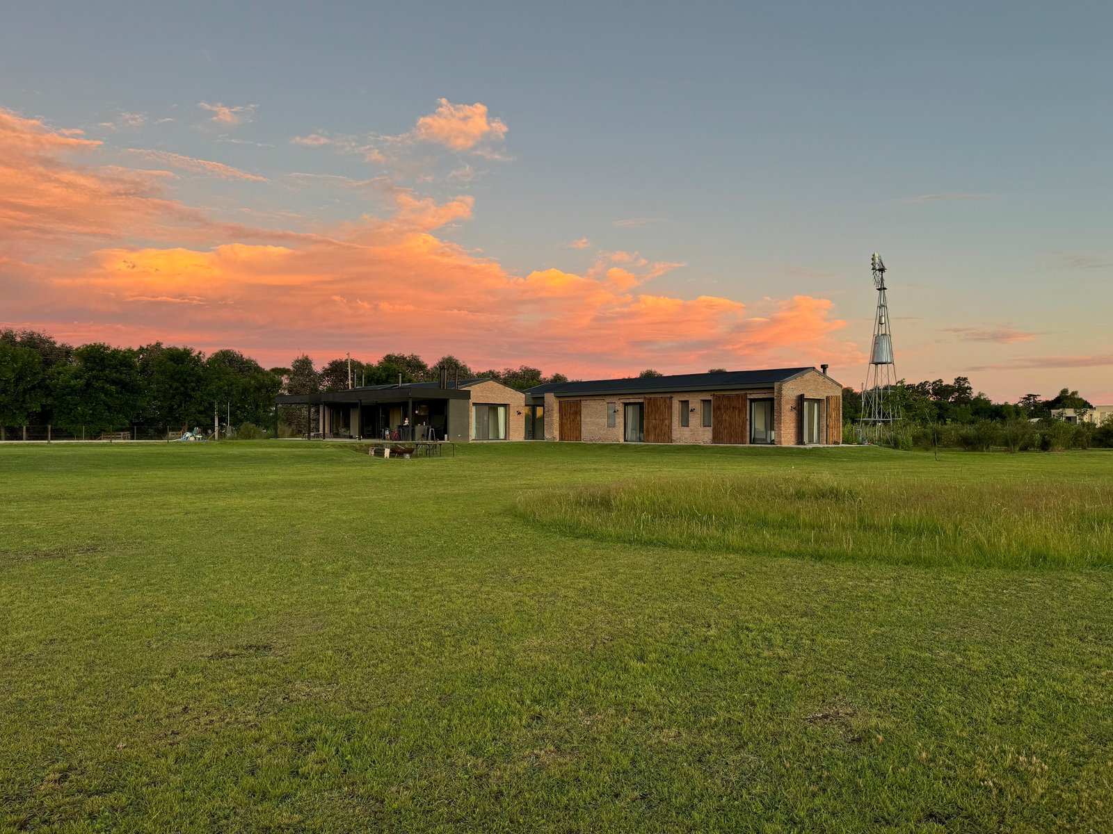 Fachada moderna em ambiente rural ao pôr do sol, com gramado e céu laranja — projeto Casa Areco, por R+B arquitectos, em San Antonio de Areco